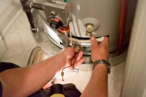 A man shining a flashlight on a hot water heater, inspecting the unit for issues in a utility room.