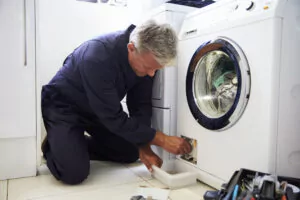 A plumber in a blue uniform repairing a household washing machine, using tools to fix the appliance in a laundry room.
